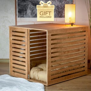 Op de afbeelding: Een lichtbruine houten dierenbench met horizontale latten en een draaideur. Binnenin de bench ligt een zacht, beige kussen. Boven de bench staat een goudkleurig icoon met een strik en de tekst GIFT INCLUDED.