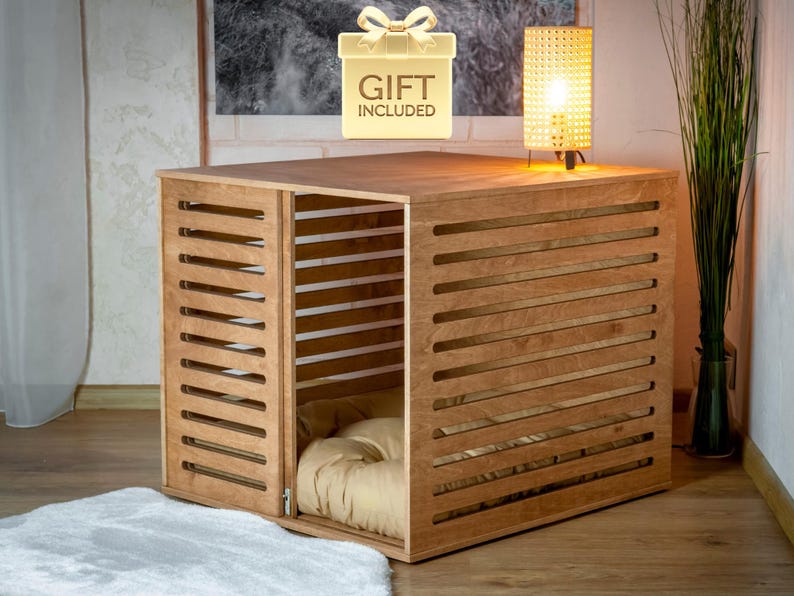 Op de afbeelding: Een lichtbruine houten dierenbench met horizontale latten en een draaideur. Binnenin de bench ligt een zacht, beige kussen. Boven de bench staat een goudkleurig icoon met een strik en de tekst GIFT INCLUDED.