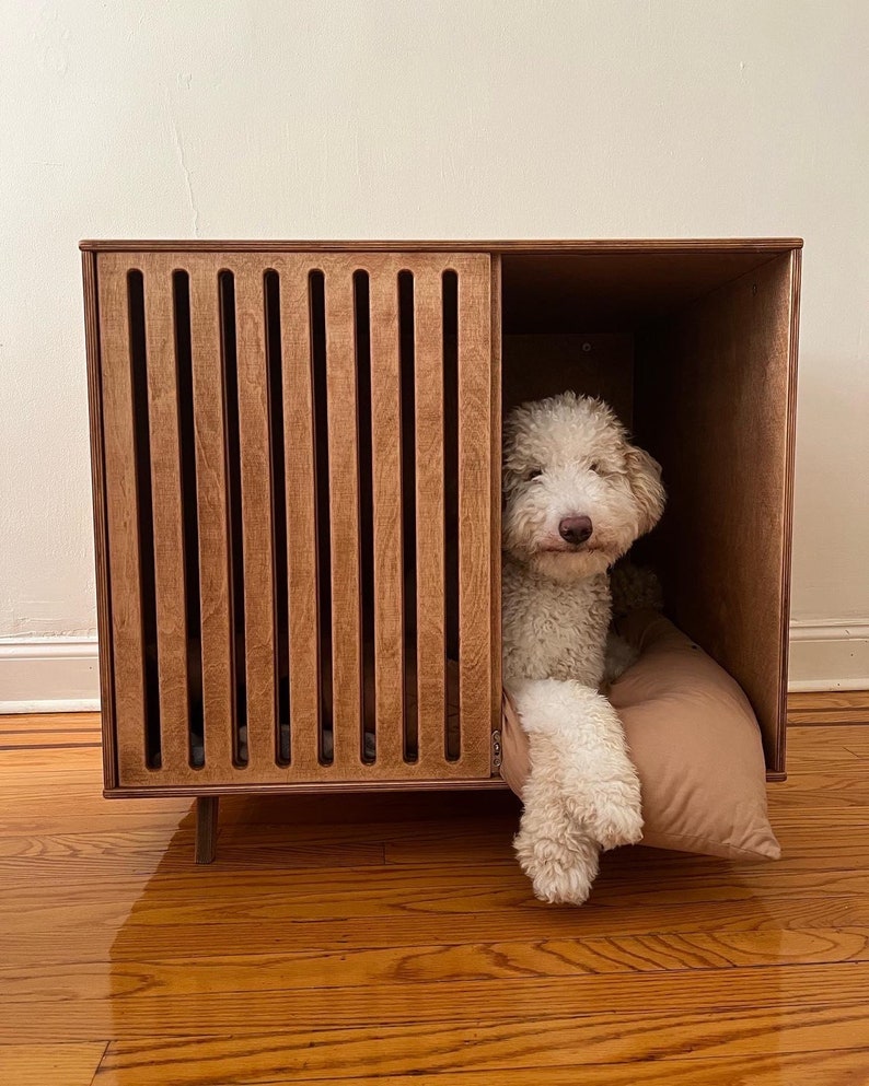 May include: A rectangular wooden dog crate with a slatted front panel and an open side section. A light-coloured, curly-haired dog is resting inside on a tan cushion, with its front paws extended outward from the open side of the wooden structure.