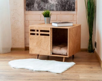 Könnte beinhalten: Ein helles Hundehaus aus Holz mit einer Schiebetür und einer rechteckigen Öffnung. Das Haus hat ein beiges Kissen im Inneren und steht auf kurzen, angewinkelten Beinen. Vor dem Haus liegt ein weißer Teppich.