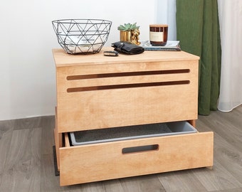 May include: A light wood cat litter box with a drawer and a slatted opening. The box has a gray litter tray inside the drawer. The box is sitting on a wood floor. There are various items on top of the box, including a black wire basket, a small potted plant, a candle, and a book.