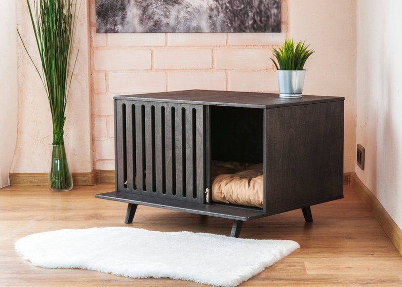 May include: A dark charcoal wooden pet crate featuring a slatted front panel, a rectangular frame, and four short angled legs. The interior contains a tan cushioned bed, and the structure is designed with a wide open side entrance.
