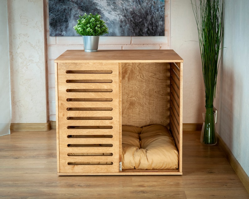 Op de afbeelding: Een lichtbruine houten dierenbench met een voorpaneel met latten en een massief bovenoppervlak. Het interieur is voorzien van een zacht, beige gecapitonneerd kussen dat de bodem van de bench bedekt voor een comfortabele rustplaats.