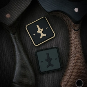 May include: Two square patches with a stylized emblem. One patch is black with a gold border and emblem, the other is dark green with a black emblem. The patches are on a textured, dark grey surface.