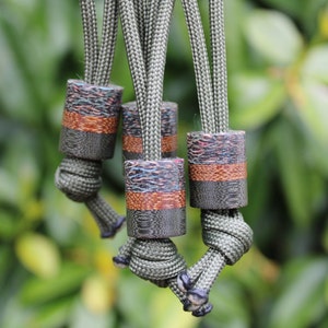 May include: Close-up of several olive green paracord lanyards with decorative cylindrical beads. The beads have a dark grey base, a copper-coloured band, and a top section with a colourful, textured pattern. The lanyards are knotted.