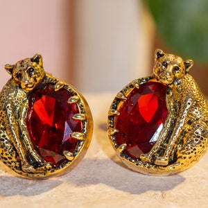 May include: A pair of gold-tone earrings featuring a detailed cheetah design holding a large red gemstone.