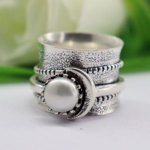 May include: A silver spinning ring with a white pearl and a crescent moon design. The ring has a textured surface and is decorated with small beads.