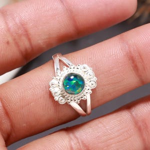 May include: A silver ring featuring a round, iridescent dark green and blue stone set in a decorative, filigree-style metal frame. The band is split into two thin sections that meet at the ornate setting, showcasing intricate metalwork details.