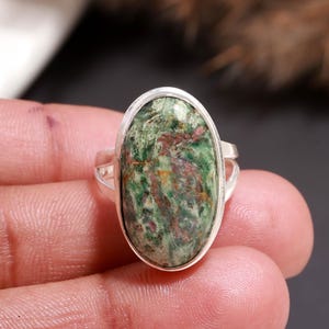 Puede incluir: Un anillo de plata con una piedra ovalada verde y roja. La piedra tiene un efecto veteado con tonos de verde, rojo y marrón. La banda del anillo se divide a ambos lados de la piedra.