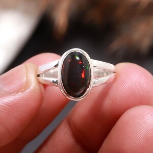 May include: A silver ring featuring a polished oval black opal gemstone set in a smooth bezel. The stone displays vibrant flashes of red and green color play. The band is crafted from polished silver and features a split shank design on both sides.
