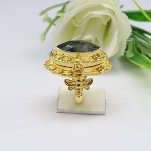 May include: A gold ring with a hinged compartment and a dragonfly design. The compartment features a dark grey gemstone.