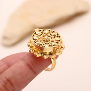 May include: A gold-toned ring with a round, ornate, hinged locket. The locket is decorated with a floral design and has a detailed, intricate pattern.