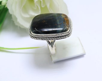May include: A silver ring with a square-shaped blue tiger's eye gemstone. The ring has a rope-like design around the edge of the setting.