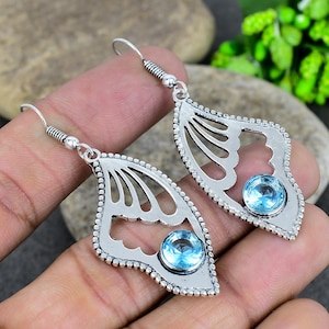 May include: Silver earrings with a leaf-shaped design, each featuring a central, round, light blue gemstone. The earrings have a decorative edge and a hook closure. The design includes openwork details.