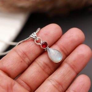 May include: A silver pendant necklace featuring a teardrop-shaped moonstone and a small, round red gemstone. The pendant is suspended from a delicate chain, showcasing the gemstones' colors and textures. The necklace is held in a hand.