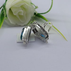 K&ouml;nnte beinhalten: Ein silberner Ring mit einem blauen Stein in der Mitte. Der Ring ist offen und hat einen Klappdeckel. Der Ring liegt auf einer wei&szlig;en Oberfl&auml;che.