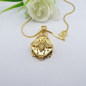 May include: A gold necklace with a bee pendant. The pendant is detailed with a textured bee design and is surrounded by a decorative border. The necklace also features a smaller bee charm.