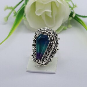 K&ouml;nnte beinhalten: Ein silberner Ring mit einem verzierten Design, das einen gro&szlig;en, facettierten, schillernden Edelstein in Form eines Sarges zeigt. Der Edelstein ist eine Mischung aus Blau, Gr&uuml;n und Lila.