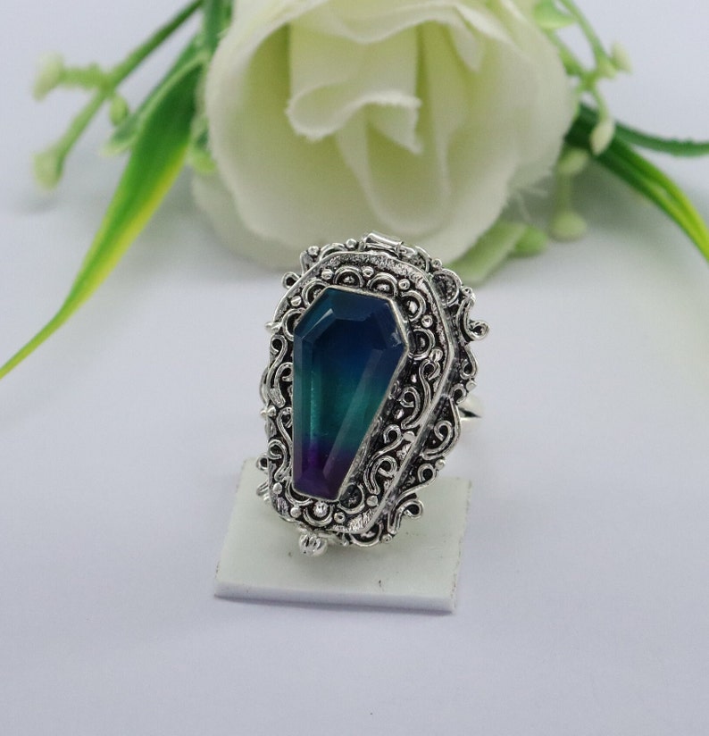 K&ouml;nnte beinhalten: Ein silberner Ring mit einem verzierten Design, das einen gro&szlig;en, facettierten, schillernden Edelstein in Form eines Sarges zeigt. Der Edelstein ist eine Mischung aus Blau, Gr&uuml;n und Lila.