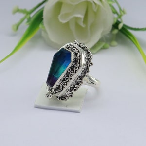 K&ouml;nnte beinhalten: Ein silberner Ring mit einem blauen und gr&uuml;nen Edelstein, der in einem Sarg-Design gefasst ist. Der Ring hat komplizierte Filigrandetails.
