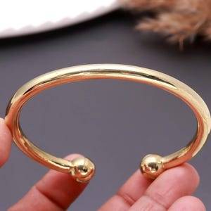Peut inclure: Un bracelet manchette en métal doré avec une finition lisse et polie. Le bracelet présente une forme circulaire ouverte, avec une tige arrondie qui se termine à chaque extrémité par un petit détail en forme de boule sphérique.