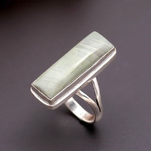 May include: A silver ring featuring a rectangular, pale green stone with subtle white linear inclusions. The stone is set in a smooth, polished silver bezel that extends into a split-shank band, showcasing a clean, metallic finish throughout the piece.