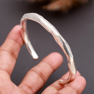 May include: A silver-colored cuff bracelet with an open design, featuring a textured, irregular, and hammered surface finish. The metal band has a curved, organic shape with varying thickness along its length, creating a distinct, uneven profile.