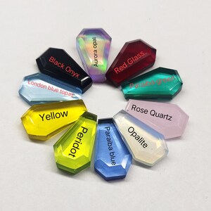 K&ouml;nnte beinhalten: Eine Sammlung von zehn Edelstein-Cabochons in verschiedenen Farben, darunter schwarzer Onyx, Londoner blauer Topas, Gelb, Aurora-Opal, Peridot, rotes Glas, Paraiba-Blau, Paraiba-Gr&uuml;n, Rosenquarz und Opalit. Jeder Cabochon ist in Form eines Sarges.