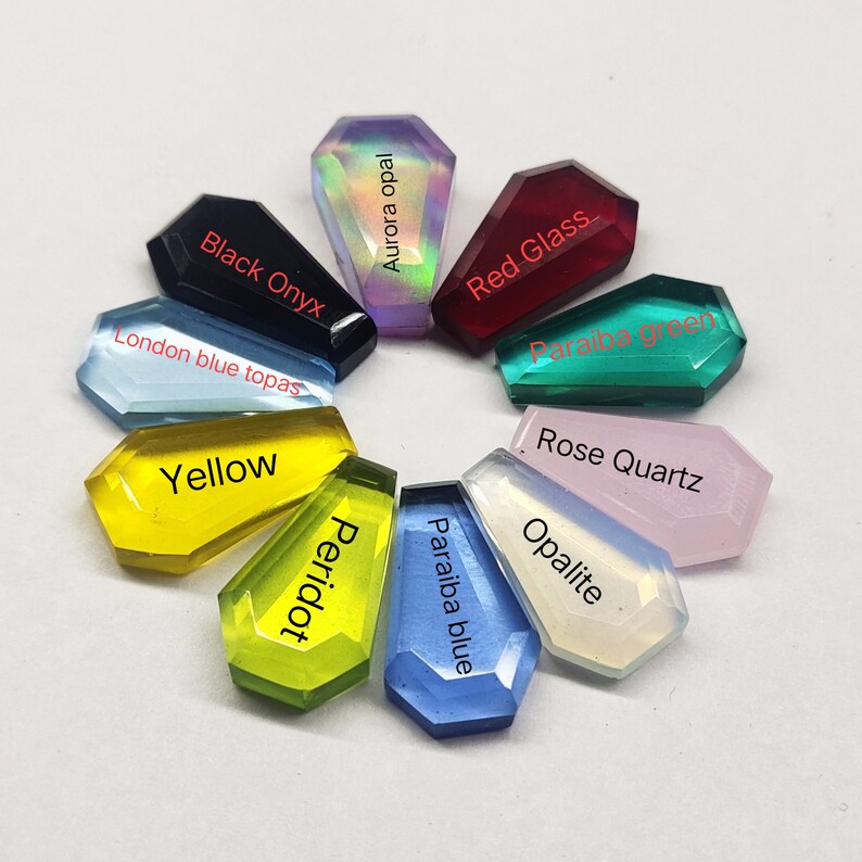 K&ouml;nnte beinhalten: Eine Sammlung von zehn Edelstein-Cabochons in verschiedenen Farben, darunter schwarzer Onyx, Londoner blauer Topas, Gelb, Aurora-Opal, Peridot, rotes Glas, Paraiba-Blau, Paraiba-Gr&uuml;n, Rosenquarz und Opalit. Jeder Cabochon ist in Form eines Sarges.