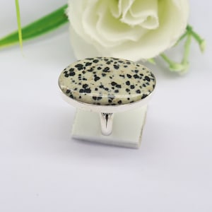 May include: Oval-shaped ring featuring a speckled, cream-colored stone with black spots, set in a silver-toned metal band. The ring is displayed on a white surface, with a blurred white flower and green stem in the background. A piece of jewelry.