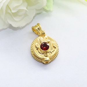 May include: A gold pendant with a red gemstone set in a floral design. The pendant has a rope-like border and a small loop at the top for attaching a chain.
