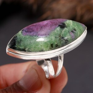 Op de afbeelding: Zilveren ring met een grote, ovale edelsteen. De edelsteen heeft paarse, groene en zwarte kleuren. De ring heeft een gepolijste zilveren band. De edelsteen is ongeveer 3 cm lang.