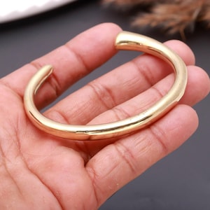 May include: A polished gold-tone cuff bracelet with a smooth, rounded surface and an open design. The metal band features a continuous, uniform thickness throughout its circular shape, showcasing a reflective, metallic finish with no additional patterns.