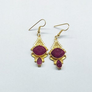 Peut inclure: Une paire de boucles d'oreilles en or avec une pierre précieuse rouge. Les boucles d'oreilles ont une forme de larme avec une plus petite pierre précieuse rouge suspendue au bas.