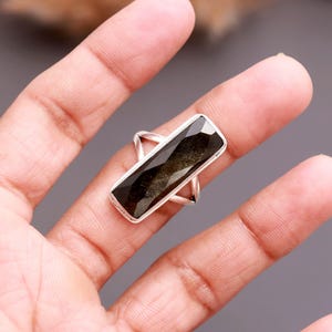 May include: A silver ring featuring a long, rectangular, faceted black and gold obsidian gemstone. The stone is set in a polished silver metal frame with a simple, thin band that wraps around the finger, showcasing a dark, reflective surface.