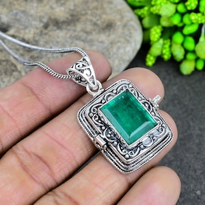 Könnte beinhalten: Ein silberner Anhänger in Form eines Medaillons mit einem verzierten Design und einem großen, smaragdgrünen Edelstein. Das Medaillon lässt sich mit einem Scharnier öffnen und schließen.
