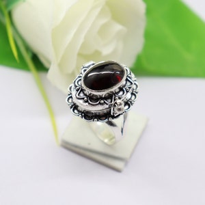 Puede incluir: Un anillo de plata con un diseño ornamentado y una piedra ovalada de color rojo oscuro. El anillo es un anillo de compartimento secreto, con una tapa con bisagras que se abre para revelar un pequeño espacio en el interior.
