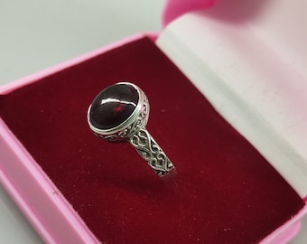 May include: A silver ring with a dark red gemstone set in a decorative bezel. The ring is displayed in a pink velvet-lined box.