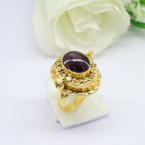 May include: A gold ring with a hinged locket featuring a purple oval gemstone. The locket is decorated with intricate floral designs.