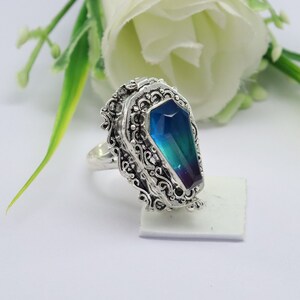 K&ouml;nnte beinhalten: Ein silberner Ring mit einem blauen und gr&uuml;nen, schillernden Stein, der in einem Sarg-Design gefasst ist. Der Ring ist mit filigranen Details verziert.