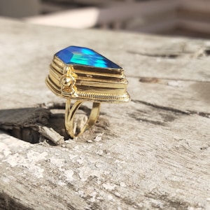 Puede incluir: Un anillo de oro con una gran piedra preciosa azul iridiscente facetada. El anillo está engastado en una banda de oro con un diseño decorativo.
