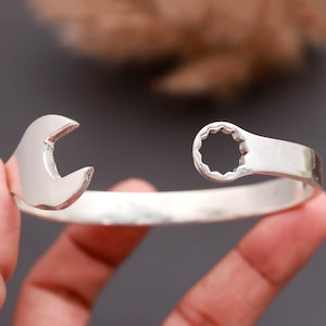Op de afbeelding: Een zilverkleurige metalen armband in de vorm van een moersleutel. Het ene uiteinde van de armband heeft een open steeksleutelkop, terwijl het andere uiteinde een gesloten ringsleutelkop heeft met een twaalfkantig intern ontwerp.