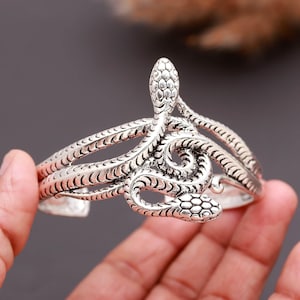 May include: A silver-toned metal cuff bracelet featuring a double-headed snake design. The textured, oxidized finish highlights the intricate scale patterns along the winding, intertwined bodies of the snakes, which meet at the center front.