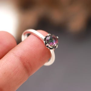 Puede incluir: Un anillo de plata con una piedra preciosa redonda e iridiscente engastada en garras. La piedra preciosa presenta tonos de rosa, morado y verde.