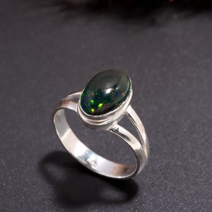 May include: A silver ring featuring an oval-shaped dark stone with iridescent green and red flecks. The band is constructed with a split shank design that connects to a smooth bezel setting holding the polished cabochon gemstone securely in place.