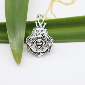 Puede incluir: Collar con colgante de plata con un diseño de abeja detallado. El colgante tiene forma de hexágono con una superficie texturizada y un contorno negro. La abeja está centrada en el colgante y tiene alas intrincadas y un cuerpo detallado.