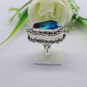 K&ouml;nnte beinhalten: Ein silberner Ring mit einem blauen und violetten Edelstein, der in einem Sarg-Design gefasst ist. Der Ring hat komplizierte Filigrandetails und ein Band, das etwas breiter ist als der Sarg.