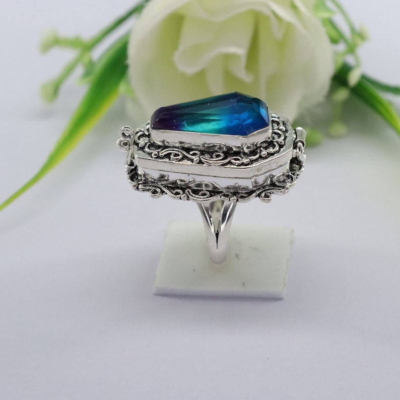 K&ouml;nnte beinhalten: Ein silberner Ring mit einem blauen und violetten Edelstein, der in einem Sarg-Design gefasst ist. Der Ring hat komplizierte Filigrandetails und ein Band, das etwas breiter ist als der Sarg.
