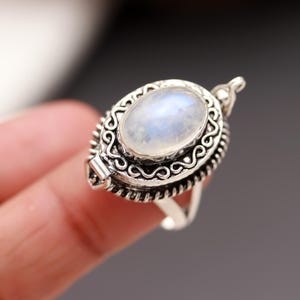 May include: A silver ring featuring an oval moonstone gemstone. The ring has an ornate, detailed design around the stone and a decorative clasp. The ring is shown on a finger.