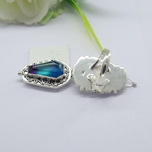 K&ouml;nnte beinhalten: Ein silberner Ring mit einem gro&szlig;en, facettierten, blauen und violetten Edelstein, der in einem detaillierten, verzierten Design gefasst ist. Der Ring wird von der Seite und von hinten gezeigt.